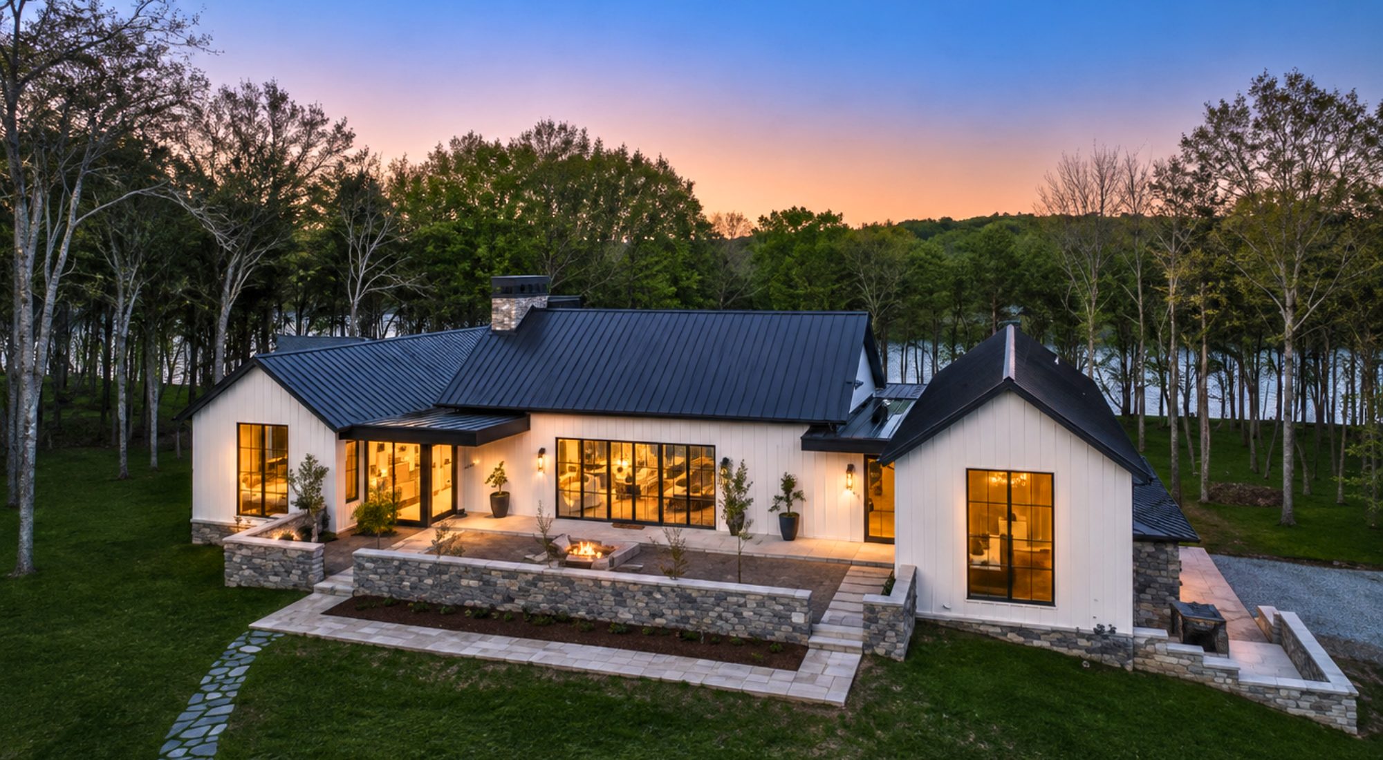 Luxury modern farmhouse at twilight with stone patio, wooded setting, and warm interior lighting | Riley King real estate portfolio